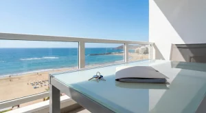 Un juego de llaves y una carpeta con un contrato de alquiler sobre una mesa en un balcón soleado con vistas a la playa de Las Canteras en Las Palmas, simbolizando la tranquilidad legal inmobiliaria en Canarias.