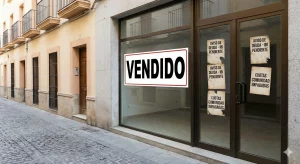 Fachada de local comercial con un cartel grande de VENDIDO en el escaparate y varios avisos de deudas de IBI y cuotas de comunidad impagadas pegados en la puerta de cristal.